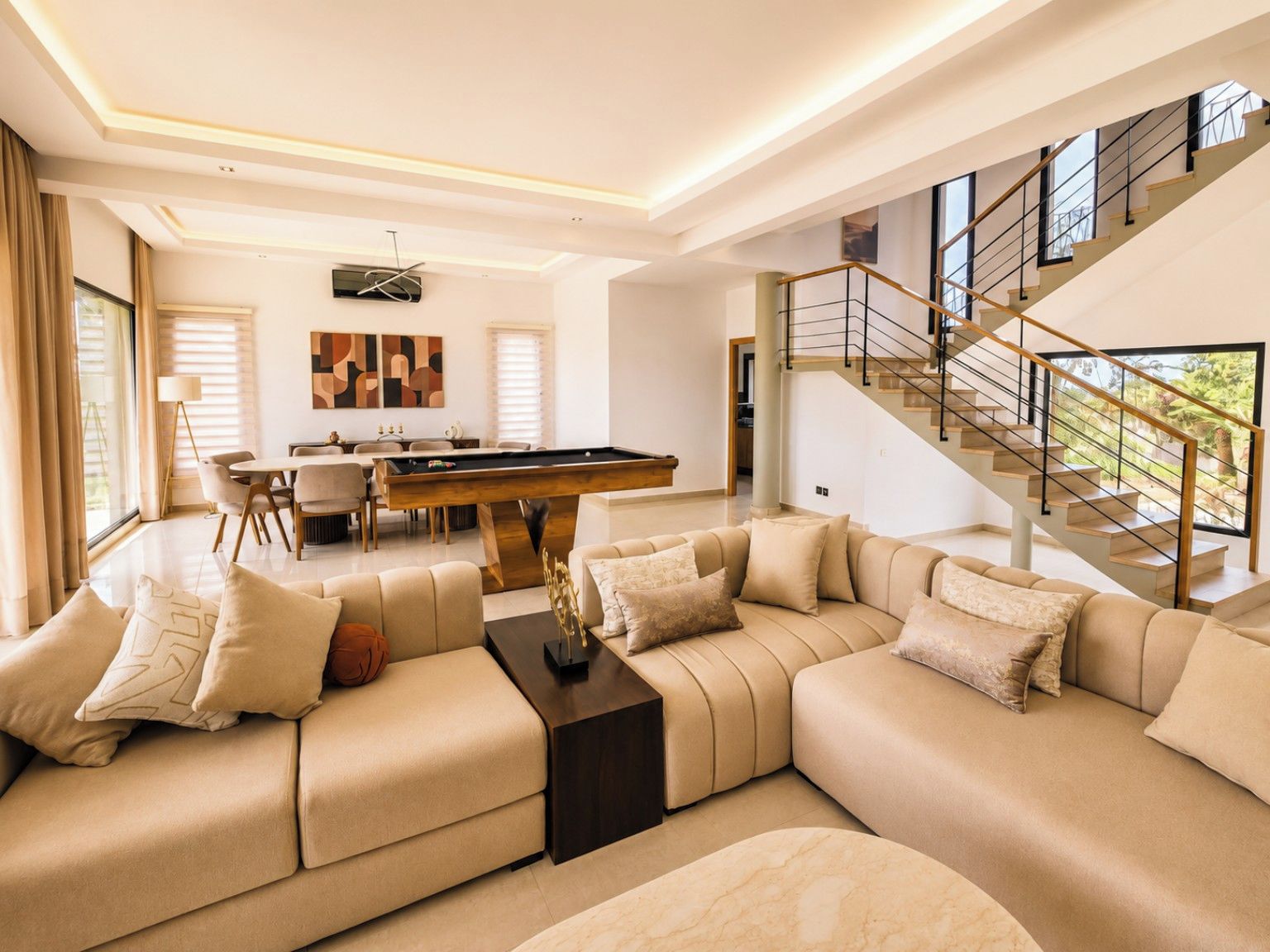A modern living room with a pool table, a sofa set, and a staircase leading to an upper level.