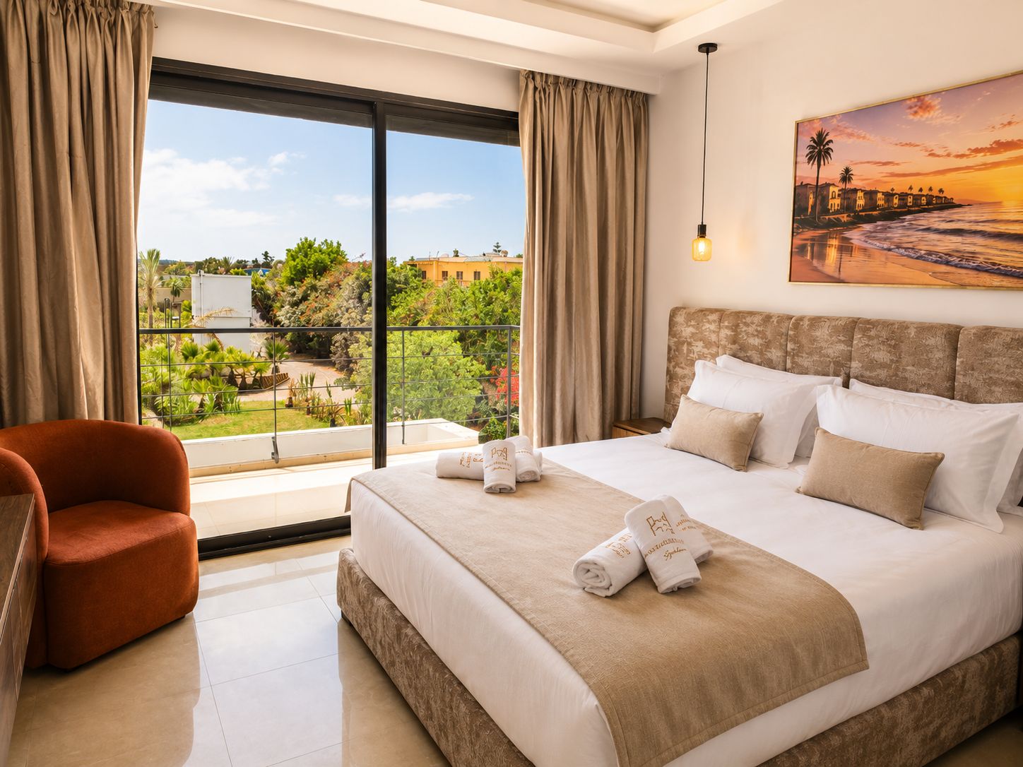 A hotel room with a large bed, a chair, a painting of a sunset, and a view of greenery and buildings outside.