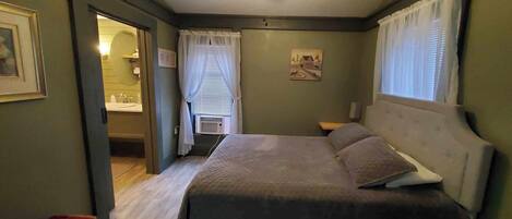 Une chambre à coucher avec un lit, une table de chevet, une fenêtre avec des rideaux, un tableau au mur et une salle de bain visible à travers une porte ouverte.