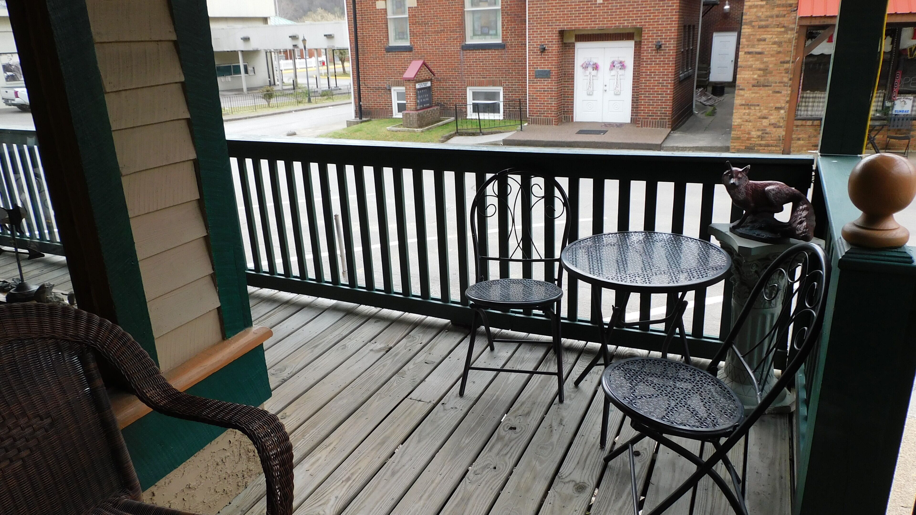 A wooden deck with metal chairs and tables, a black railing, and a view of a brick building with a white door.