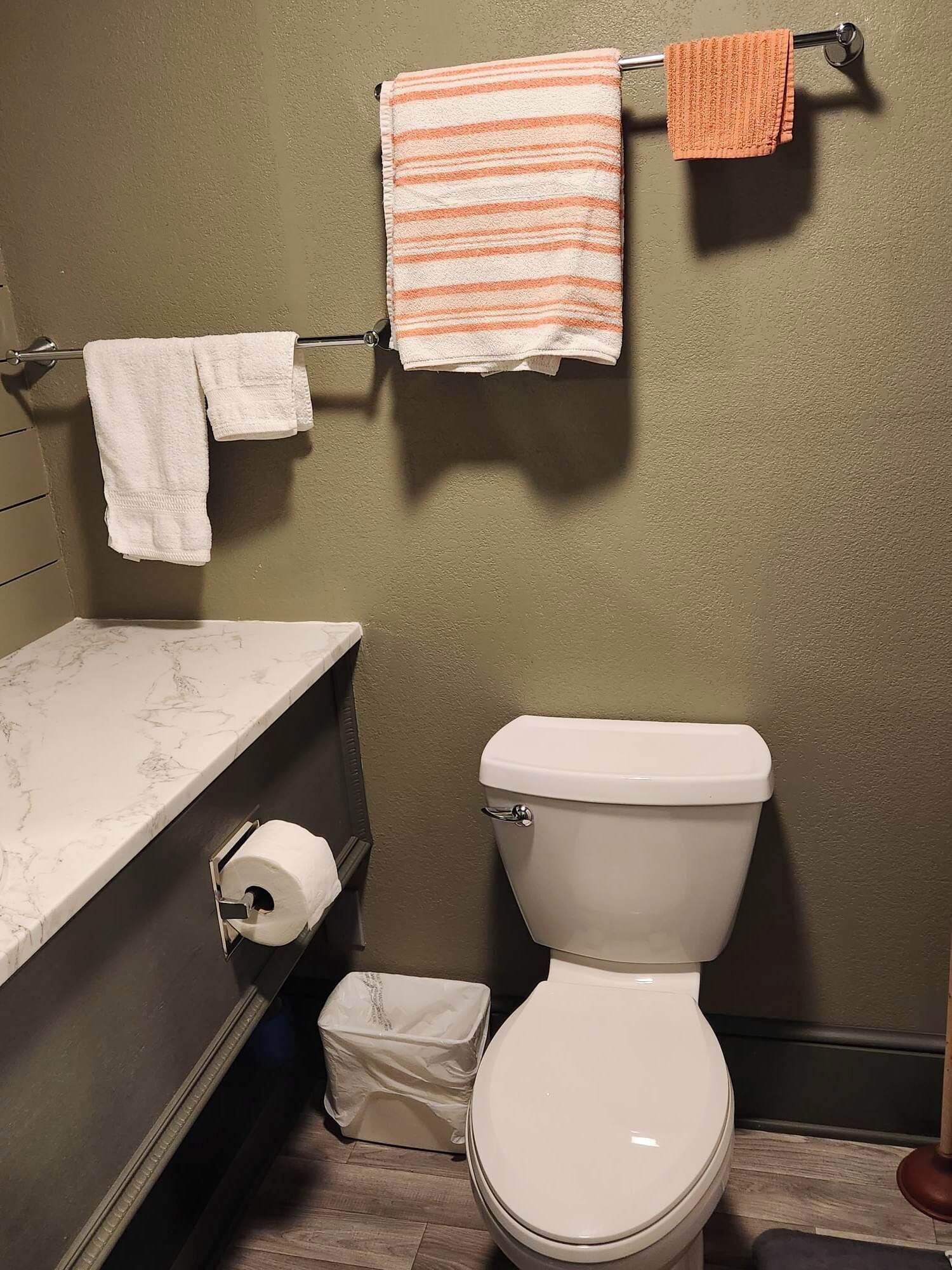 Un baño con inodoro, encimera de mármol, toallero con dos toallas y dispensador de papel higiénico.