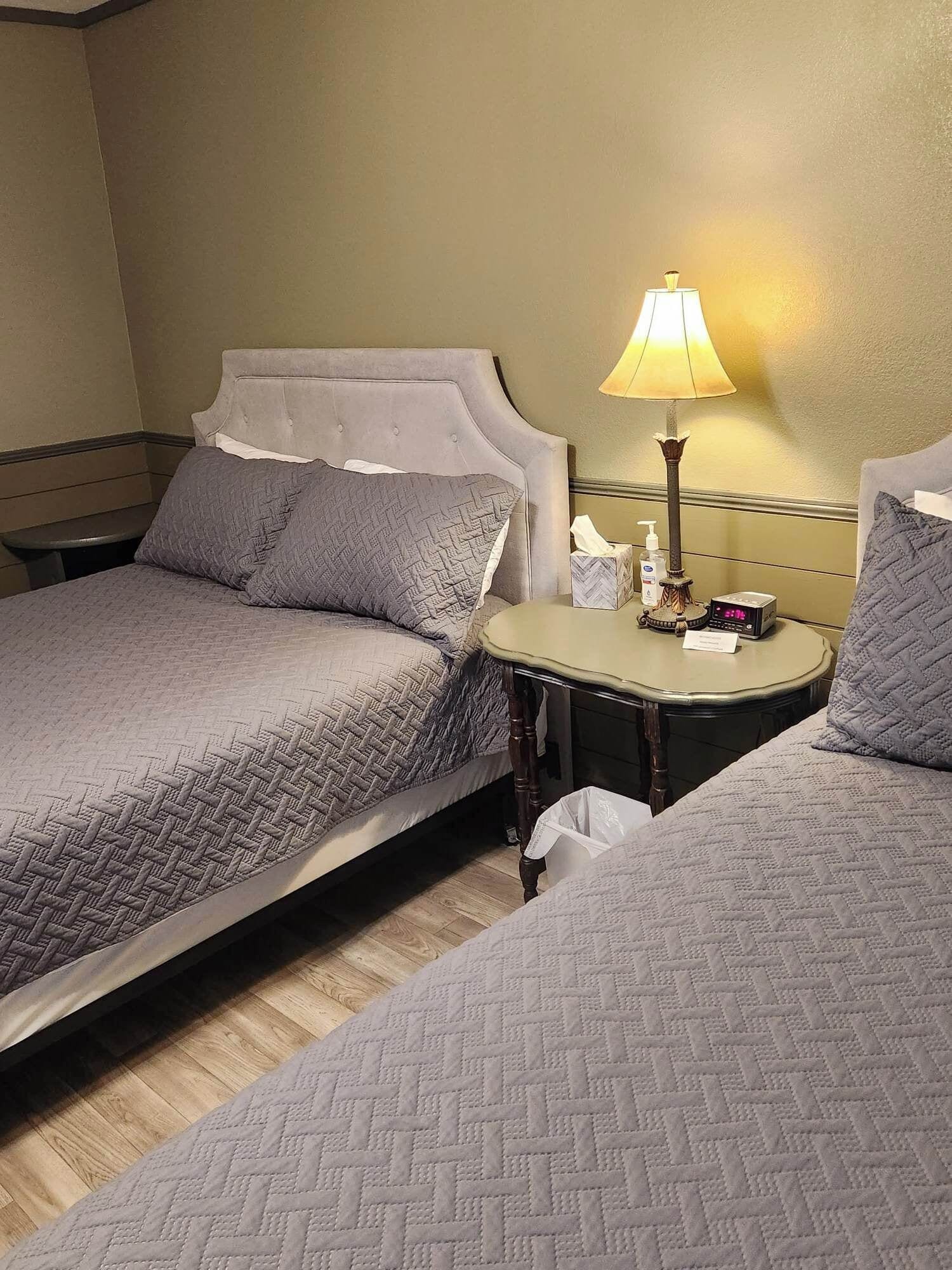 Une chambre d’hôtel avec deux lits, une table de chevet avec une lampe, une petite table de nuit avec une horloge et une poubelle.