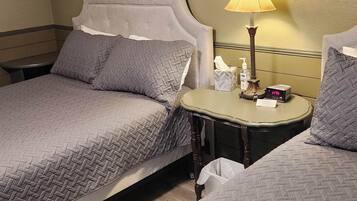Une chambre d’hôtel avec deux lits, une table de chevet avec une lampe, une petite table de nuit avec une horloge et une poubelle.