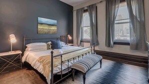 A bedroom with a bed, bedside lamps, a bench, a painting, and a window with curtains. - Casa Hotels: Chic Industrial 2nd Floor Suite (Toronto)