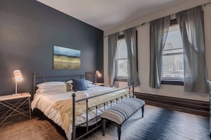 Ein Schlafzimmer mit einem Bett, Nachttischlampen, einer Bank, einem Gemälde und einem Fenster mit Vorhängen.