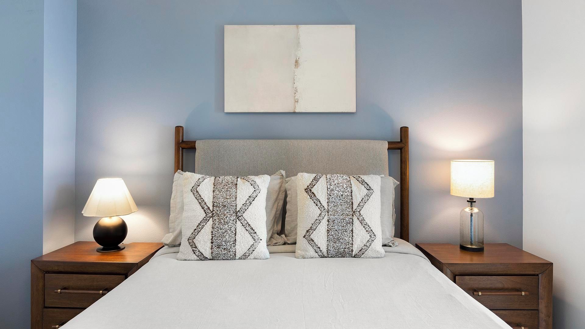 A neatly made bed with two patterned pillows, a bedside lamp, and a wooden nightstand with a drawer.