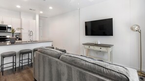 A modern living space with a sofa, a kitchen area, a flat-screen TV, and a bar stool. - Landing | Stunning 2BD, Gym (North Las Vegas)