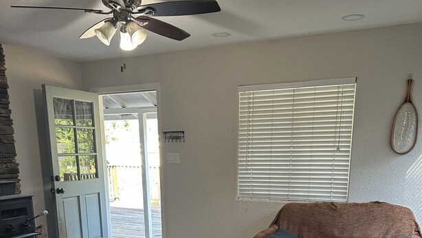 A living room with a stone fireplace, a ceiling fan, a brown sofa, a wooden rocking chair, and a large window with blinds.