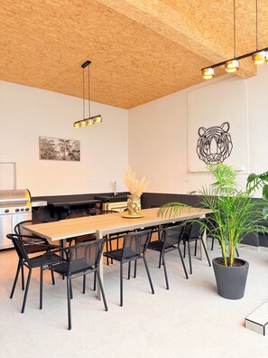 Eine moderne Küche mit Holzbalkendecke, einem langen Esstisch, schwarzen Stühlen, einem Grill und einem Topf mit Pflanze.
