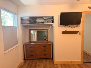 Ein Zimmer mit einem Holzkasten, einem Spiegel, einem an der Wand befestigten Fernseher und einem Fenster mit Jalousien.