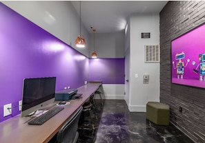 Ein moderner Büroraum mit violetten Wänden, einem langen Tisch mit Computern und einer Wand als Ziegelakzent.