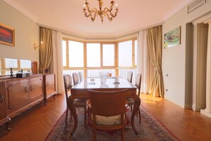 Uma sala de jantar com uma mesa grande, cadeiras, um candelabro e vista através de grandes janelas.