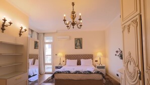 A bedroom with a tufted headboard, a large bed with white bedding, two bedside lamps, a chandelier, a wardrobe with decorative handles, and a door with an ornate design. - Nile front view & pyramids too.3 master BR+maid room.Large classy fully equipped (Cairo Governorate)