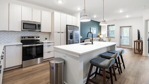 Cocina moderna con armarios blancos, electrodomésticos de acero inoxidable, una isla central con taburetes y luminarias colgantes.