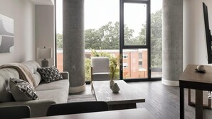 Ein modernes Wohnzimmer mit Sofa, Tisch und Stuhl, ein großes Fenster mit Blick auf Gebäude und Bäume, und einer Lampe.