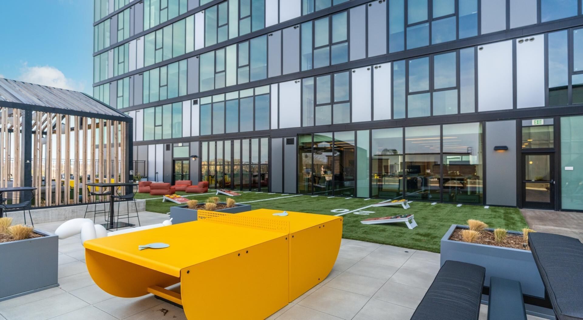 A modern building with large windows, a courtyard with outdoor seating, and a yellow table.