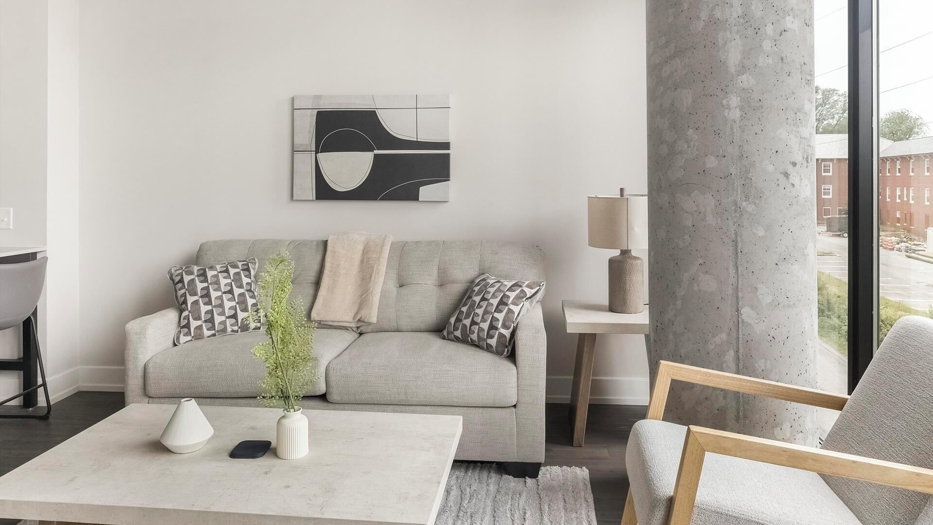 Un salón moderno con un sofá gris, cojines estampados, una mesa de centro blanca, un jarrón con follaje y una obra de arte minimalista en la pared.