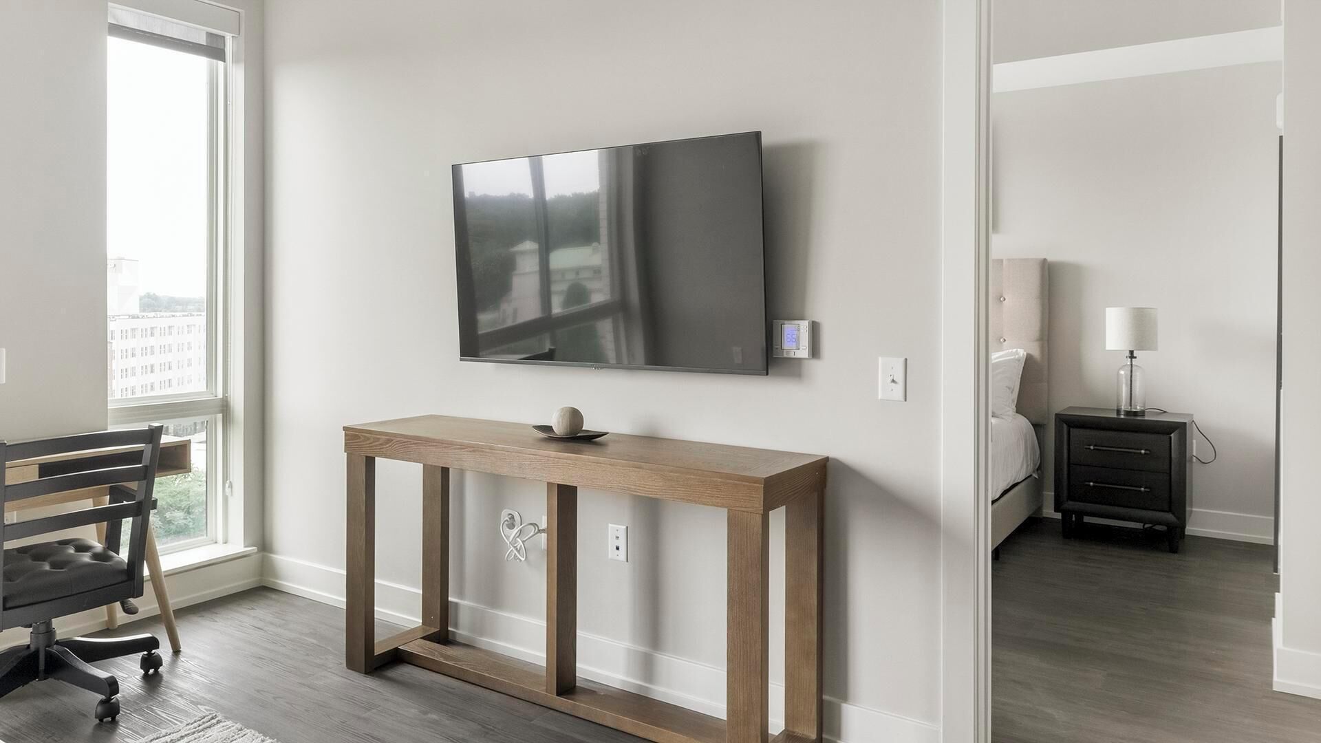 Habitación moderna con una televisión de pantalla plana grande montada en la pared, una mesa consola de madera con un adorno, una silla de oficina negra y una mesita de noche con una lámpara.