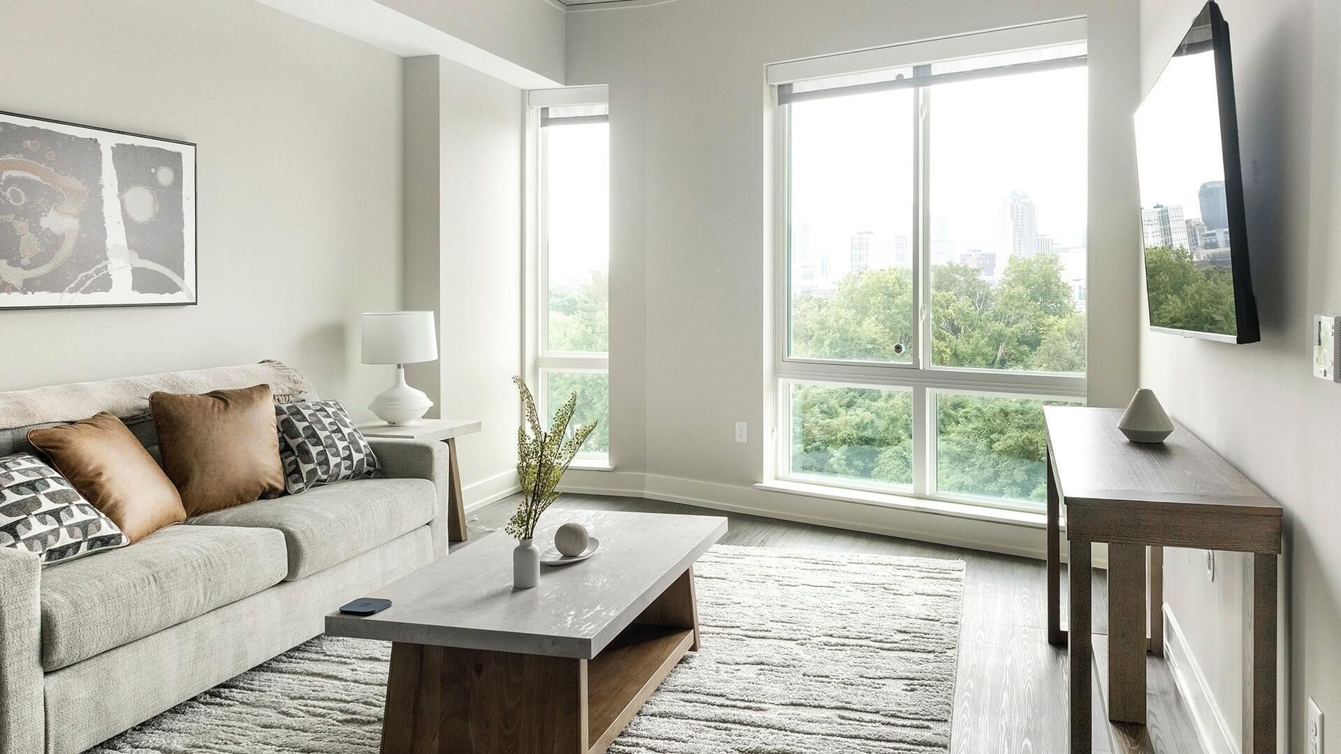 A modern living room with a grey sofa, a coffee table, and a large window offering a city view.
