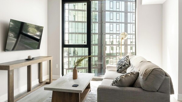 Ein modernes Wohnzimmer mit einer Couch, einem Couchtisch, einem an der Wand montierten Fernseher und großen Fenstern mit Blick auf Gebäude.