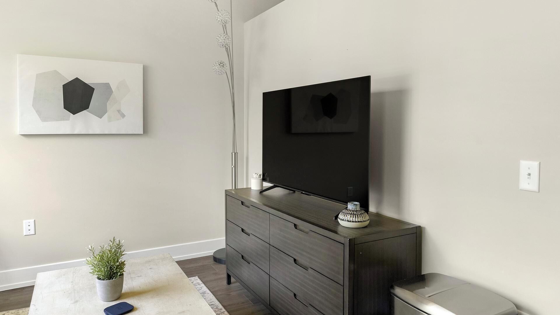 Una sala de estar moderna con un televisor de pantalla plana montado en un mueble de madera oscura, un cuadro abstracto minimalista en la pared, una planta en maceta y un pequeño florero sobre el mueble.