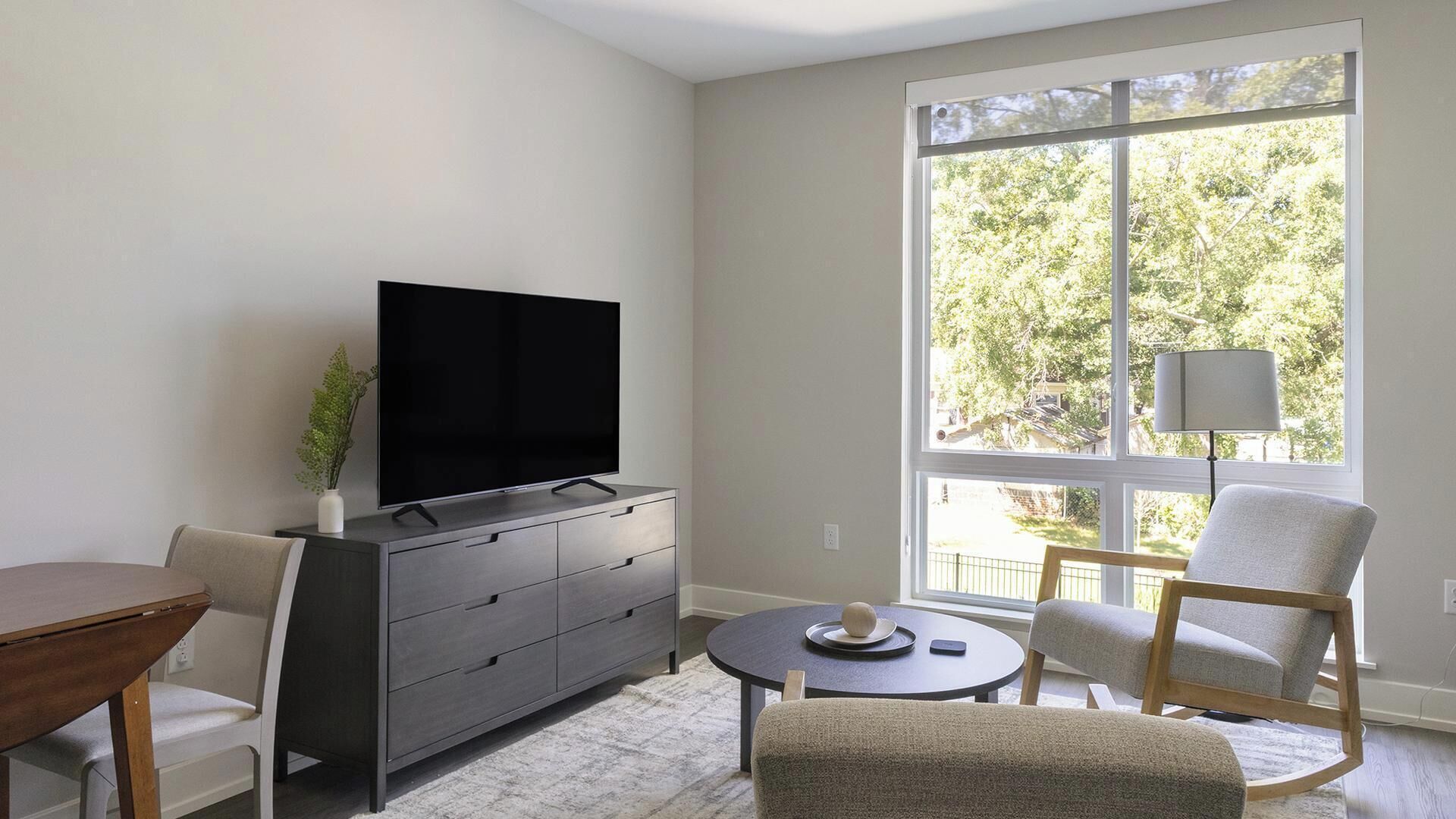 Una sala de estar con una televisión de pantalla plana sobre un mueble bajo, una mesa de centro redonda, una silla y un ventanal con vistas a árboles.