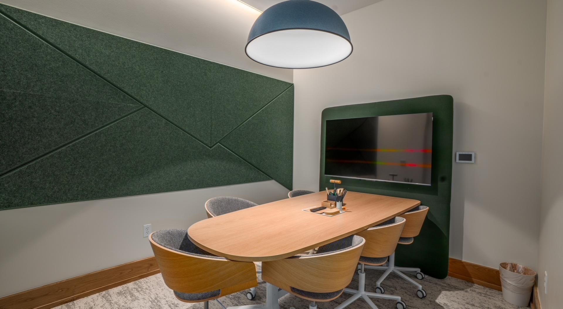 Een vergaderruimte met een houten tafel, vier stoelen, een groot scherm en een groene accentmuur.