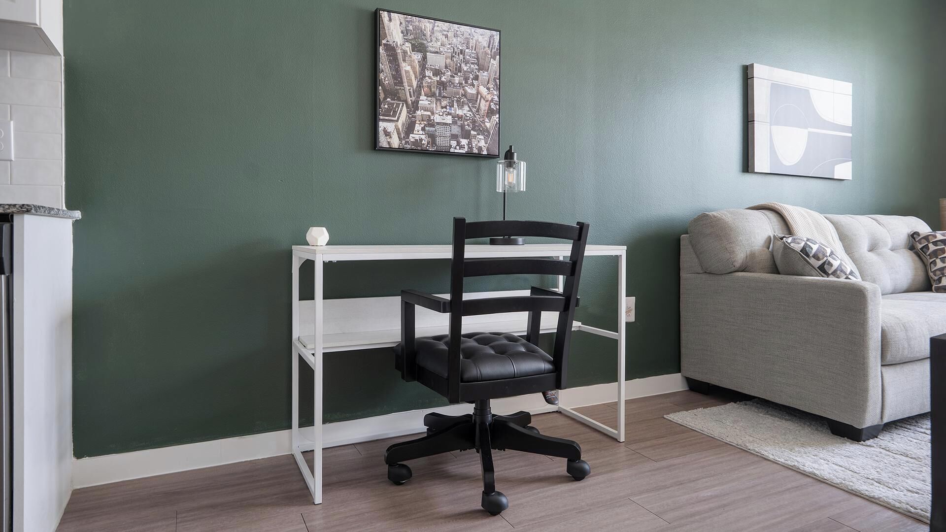 A room with a white desk, a black office chair, a grey sofa, and a green wall.