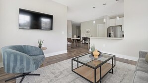 A modern living room with a flat-screen TV, a blue armchair, a glass coffee table, and a kitchen area in the background. - Landing | Stylish 2BD, Game Room, Gym (Charlotte)