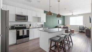 A modern kitchen with white cabinets, a stainless steel refrigerator, a microwave, and a stove. There is a large island with bar stools. - Landing | Chic 1BD, Gym, Clubhouse (Kyle)