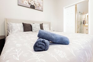 Ein ordentlich bezogenes Bett mit einer blumigen Tagesdecke, ein aufgerolltes Handtuch und ein gerahmtes Blumenbild über dem Bett.