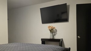 A bedroom with a bed, a flat-screen TV, a small table with a vase, and a door.