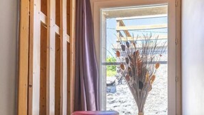 Quarto com piso de madeira, um banquinho vermelho, um vaso com plantas secas e vista pela janela.
