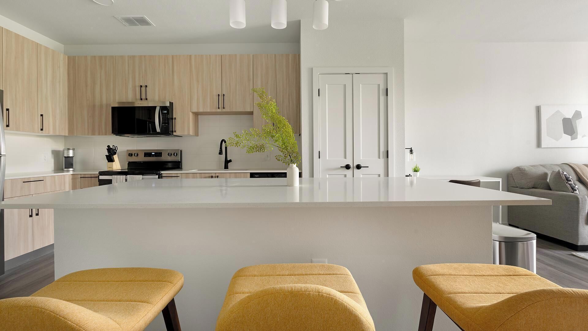 Una cocina moderna con una isla central, armarios de madera, un microondas y un lavabo. Hay tres taburetes amarillos frente a la isla.