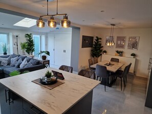 A modern kitchen with a central island, dining area with chairs, and a sofa with cushions. - Dommar Guest House Luxury & Comfort (Saint Mellons)