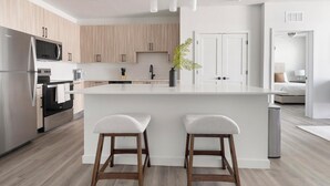A modern kitchen with a central island, wooden cabinets, stainless steel appliances, and a view into a bedroom. - Landing | Spectacular 2BD, Gym, Clubhouse (North Fort Myers)