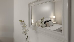 Una camera d'albergo con uno specchio grande, un letto con biancheria bianca, una lampada da comodino e un vaso con fiori bianchi.