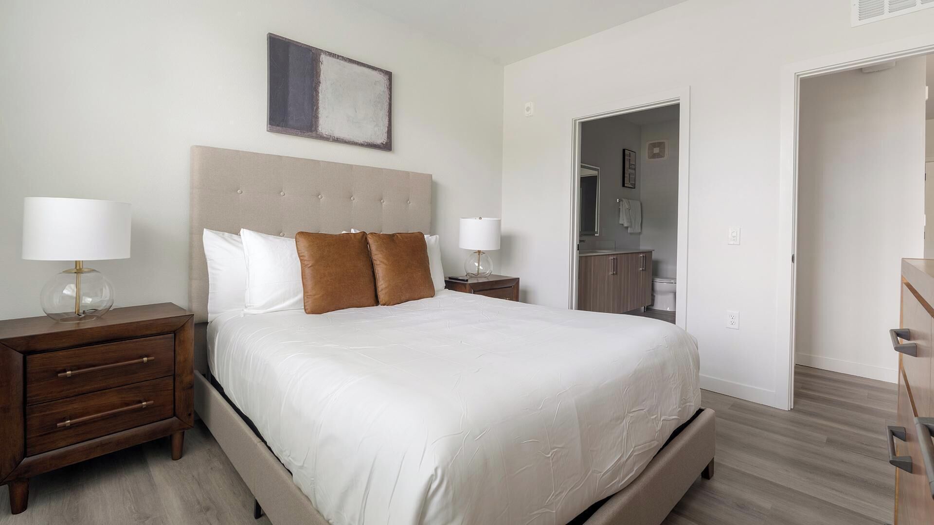 A bedroom with a bed, two bedside tables, a nightstand, a headboard, a wall-mounted artwork, and an open door leading to a bathroom.