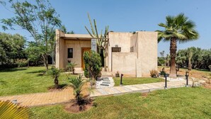 Un edificio moderno con tetto piatto, ingresso pavimentato e una piccola area giardino con palme e arbusti.
