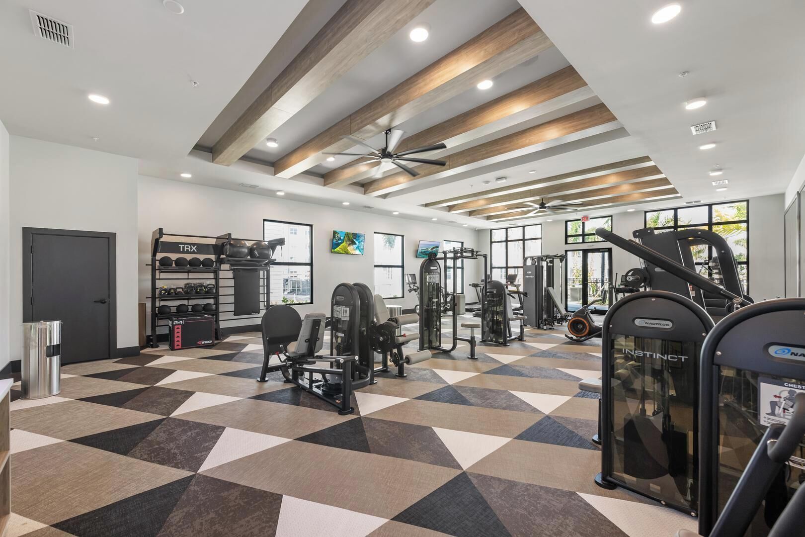 Uma academia com diversos equipamentos de ginástica, um ventilador de teto e piso xadrez.