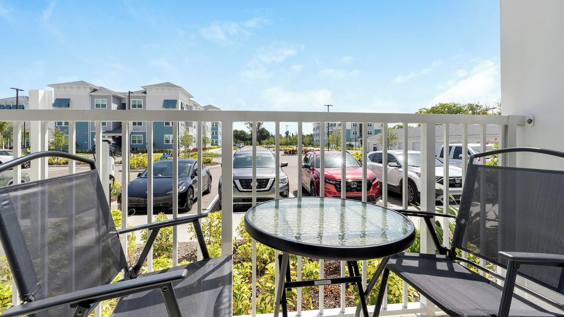 Un balcone con tavolo e sedie, affaccia su un'area di parcheggio con diverse auto e edifici sullo sfondo.