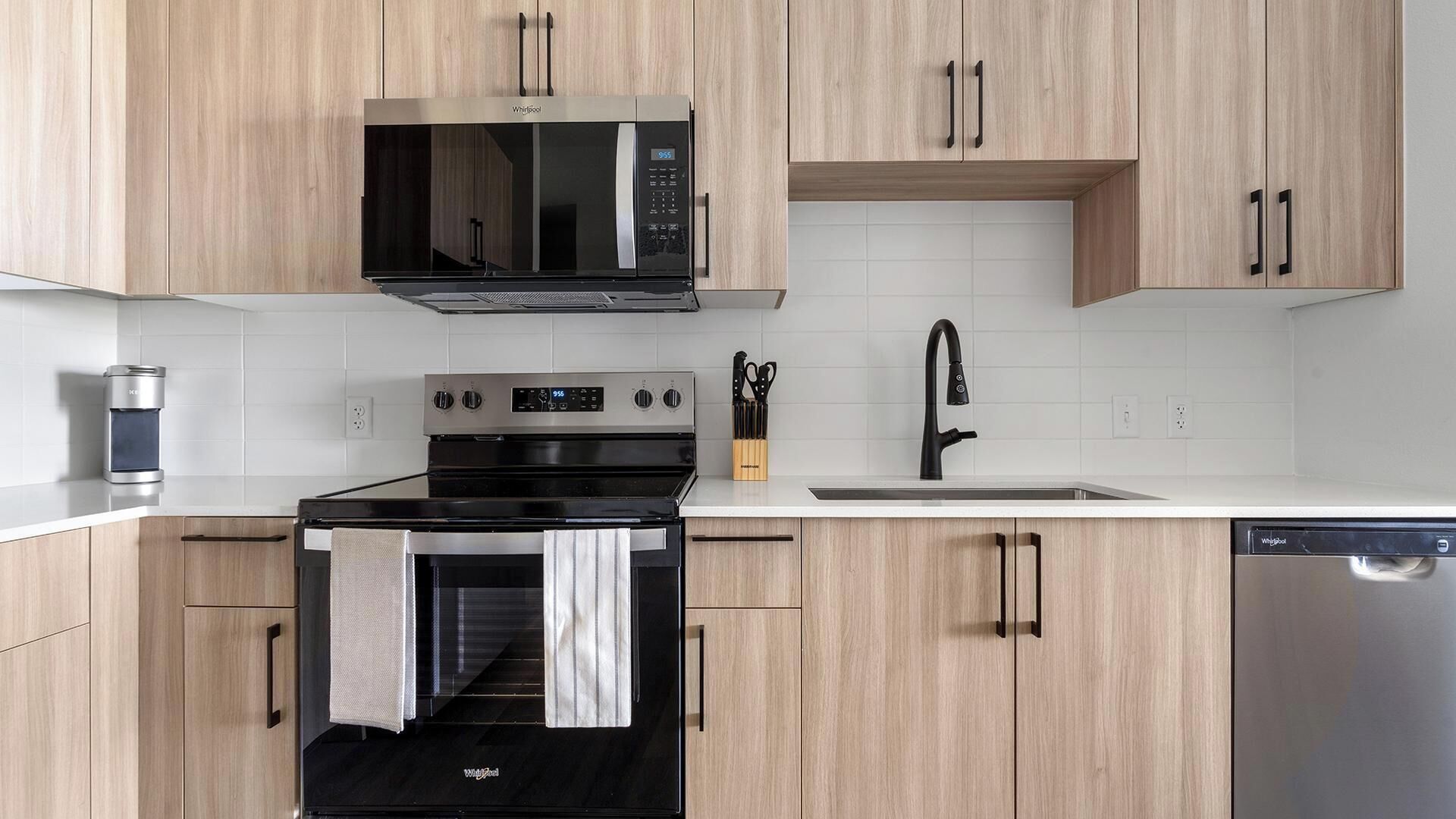 A modern kitchen with a built-in microwave, oven, and dishwasher, wooden cabinets, and a white tiled backsplash.