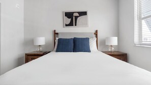 Habitación de hotel con cama, dos mesitas de noche con lámparas, una obra de arte en la pared y una ventana con persianas.