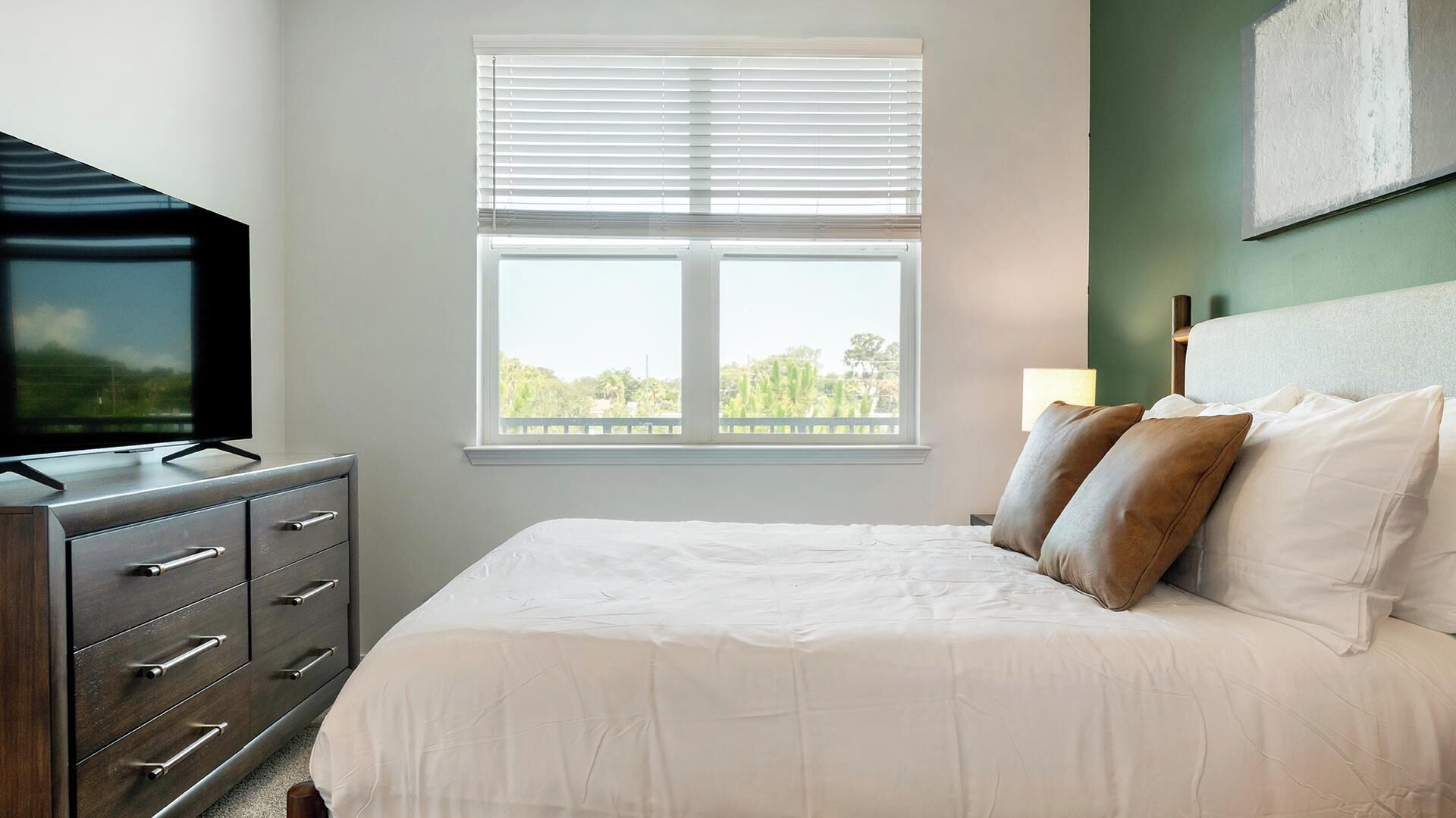 A bedroom with a bed, a dresser, a television, and a window with blinds.