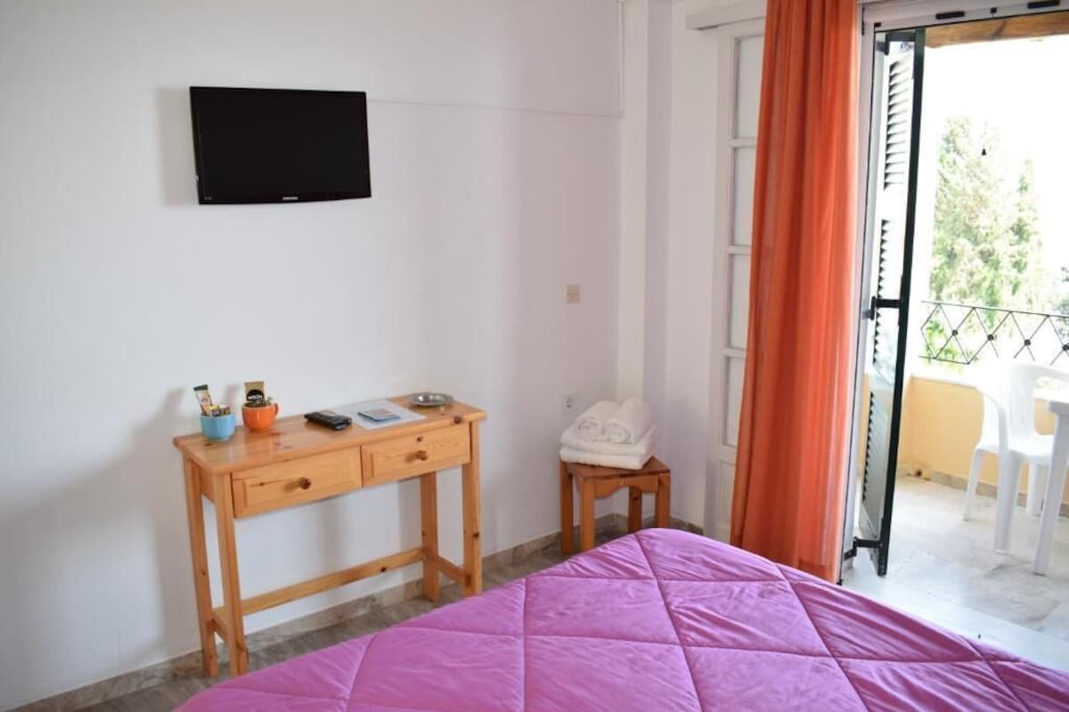 Ein Schlafzimmer mit Bett, Schreibtisch inklusive Fernseher, einem kleinen Tisch mit Handtüchern und einem Balkon mit Stühlen.