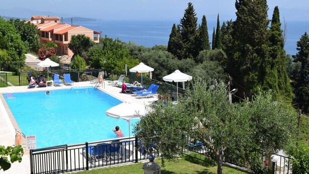 A swimming pool with lounge chairs and umbrellas, surrounded by greenery and overlooking a body of water. - Rising Sun Apartments Studios (Corfu)