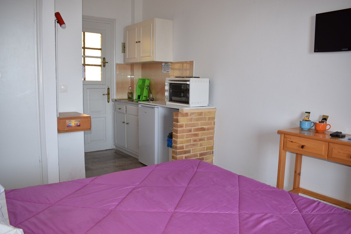 Ein Schlafzimmer mit einer violetten Bettdecke, einem Holznachttisch mit Tasse und Fernbedienung, einem kleinen Tisch mit einer Mikrowelle und einer Tür mit Fenster.