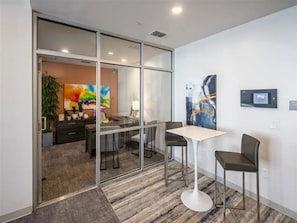 A modern office space with a glass partition, a white table, two chairs, a desk with a computer, and a painting on the wall. - Landing | Stylish 2BD, Clubhouse (Tarpon Springs)