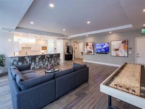 A modern lounge area with a sofa, a ping pong table, and a kitchen in the background. - Landing | Stylish 2BD, Clubhouse (Tarpon Springs)