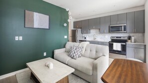 A modern living room with a sofa, a coffee table, and a dining table. There is a kitchen area with grey cabinets and appliances. - Landing | Spectacular 1BD, Courtyard, Pool (Richmond)